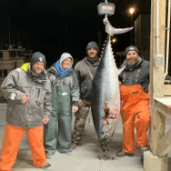 Four people posing with large fish hanging on a scale in a dimly lit area.